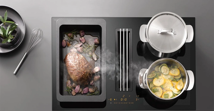 Ein Braten mit Zwiebeln und Kräutern schmort in einer Pfanne auf einem modernen Kochfeld mit Dampfabzug. Daneben kochen zwei Töpfe, einer mit Deckel, der andere mit Gemüse. Auf der linken Seite steht ein Metallbesen neben einem Teller mit frischen Kräutern.