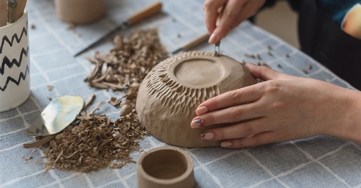 Frau verziert Keramikschüssel in Rohzustand mit einem Messer am unteren Rand. Auf dem Tisch sind Tonreste, eine fertige Schale und weiteres Werkzeug zu sehen.