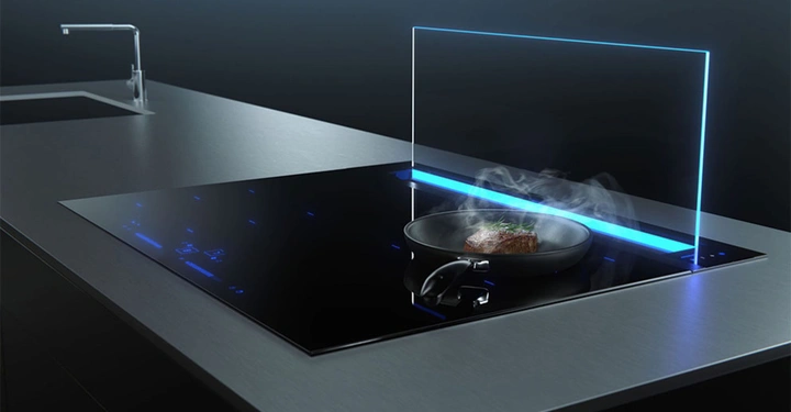 Pfanne brät ein Stück Fleisch auf einem modernen Induktionskochfeld in einer minimalistischen Küche mit Edelstahl-Arbeitsplatten und einem Wasserhahn neben einem Spülbecken im Hintergrund.