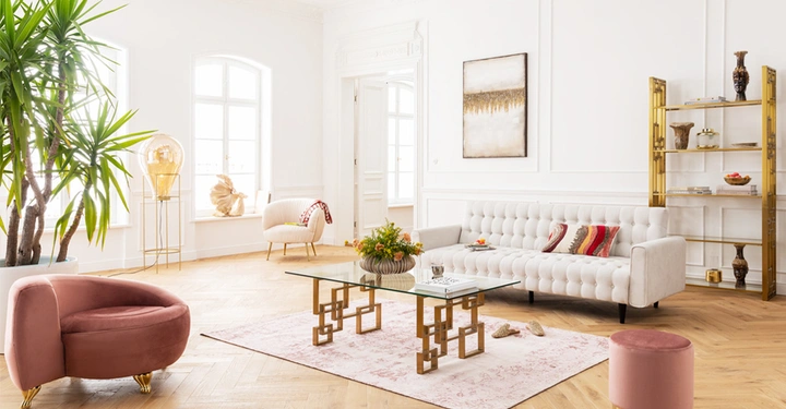 Galerie-Bild 6: Ein weißes, lichtdurchflutetes Wohnzimmer mit einer weißen Couch auf einem rosa Teppich, flankiert von einem goldenen Regal. Eine große Zimmerpflanze und ein rosa Sessel ergänzen die Einrichtung.