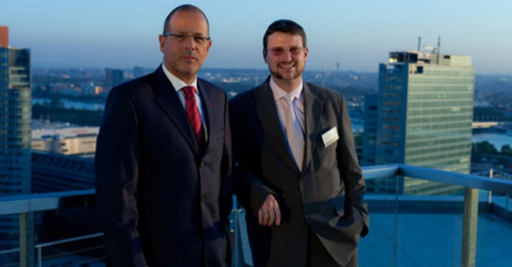 Galerie-Bild 1: Zwei Männer in Anzügen stehen auf einem Balkon eines Hochhauses. Einer lächelt leicht der andere schaut ernst. Im Hintergrund ist eine Stadtlandschaft bei Sonnenuntergang zu sehen.