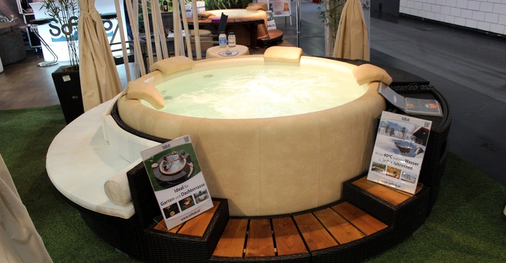 Galerie-Bild 24: Ein runder Whirlpool mit Holzstufen wird auf einer mit Gras bedeckten Ausstellungsfläche präsentiert. Plakate bewerben den Whirlpool als ideal für Garten- und Dachterrassen www.softub.at.