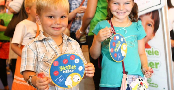 Zwei Kinder halten bunte runde Schilder in den Händen und lächeln in die Kamera auf einer Messe Veranstaltung im Hintergrund Menschen und ein großer Comic-Ausschnitt auf einem Werbeplakat Text: "Herbst Messe".