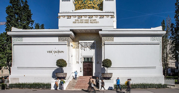 Weißes Gebäude mit kunstvollen Verzierungen ist von Bäumen umgeben und befindet sich in einer städtischen Umgebung. Text enthält VER SACRUM DER ZEIT IHRE KUNST DER KUNST IHRE FREIHEIT.