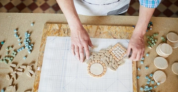 Person legt auf einem Blatt Papier nach einem vorgezeichneten Muster Mosaiksteinchen auf, das Blatt Papier liegt auf einer rechteckigen Grobspanplatte