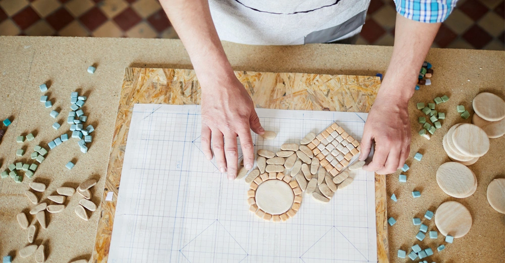 Person legt auf einem Blatt Papier nach einem vorgezeichneten Muster Mosaiksteinchen auf, das Blatt Papier liegt auf einer rechteckigen Grobspanplatte