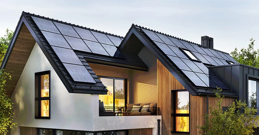 Großes Einfamilienhaus in Abenddämmerung, von innen beleuchtet mit großen Photovoltaik-Paneelen am Dach. Im Vordergrund Sträucher und Bäume.