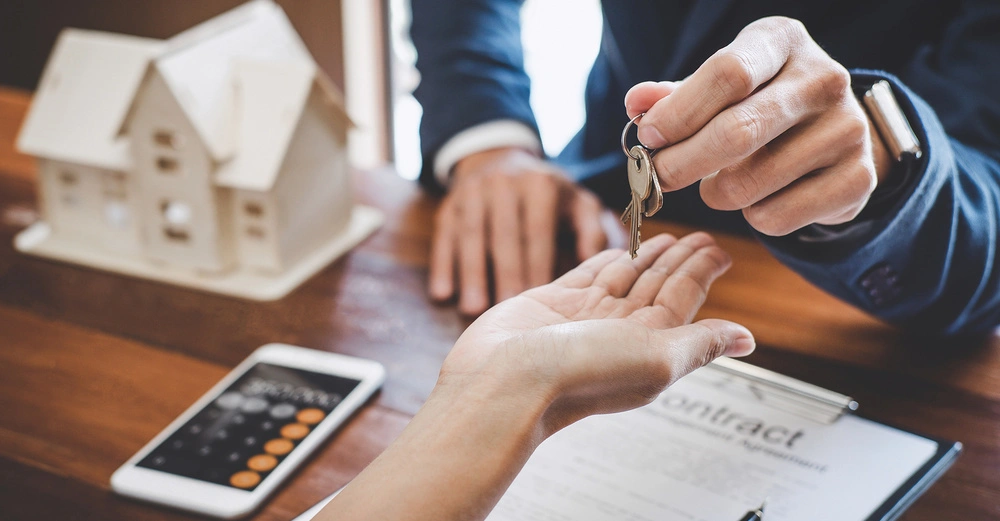 Ein Schlüssel wird von einer Person an eine andere übergeben im Hintergrund ein kleines Hausmodell auf einem Holztisch daneben ein Smartphone und ein Papier mit dem Wort "contract"