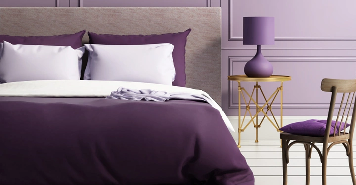 Boxspringbett mit lila Tagesdecke, daneben Nachtkästchen mit violetter Stehlampe, violetter Samtstuhl, fliederfarbene Wand. 