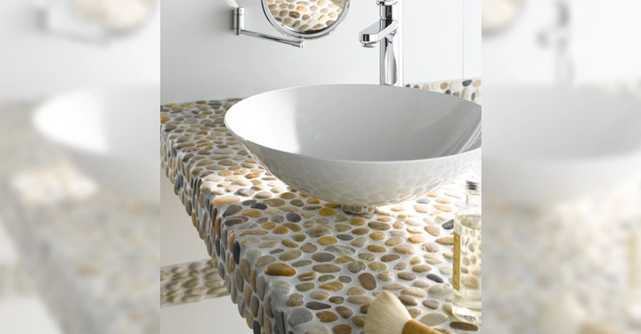 Galerie-Bild 2: Waschbecken stehend auf einer Arbeitsfläche aus Kieseln ruht in einem modernen Badezimmer mit Spiegel Wandhahn Seifenspender und Pinsel in einem hell erleuchteten Raum.