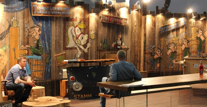 Galerie-Bild 8: Zwei Männer sitzen an Holztischen in einem Raum mit bemalten Holzwänden und Holzmöbeln STAMM DESIGN Tischmanufaktur ist auf Schildern zu lesen Ein Schild sagt Horske Werbung von STAMM Hängeleuchten beleuchten den Raum