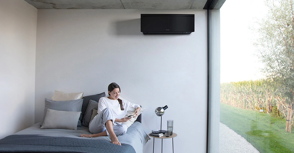 Junge Frau sitzt in einem modernen Schlafzimmer auf einem Doppelbett und liest, rechts über ihr an der Wand ist eine schwarze Luft-Luft-Wärmepumpe montiert