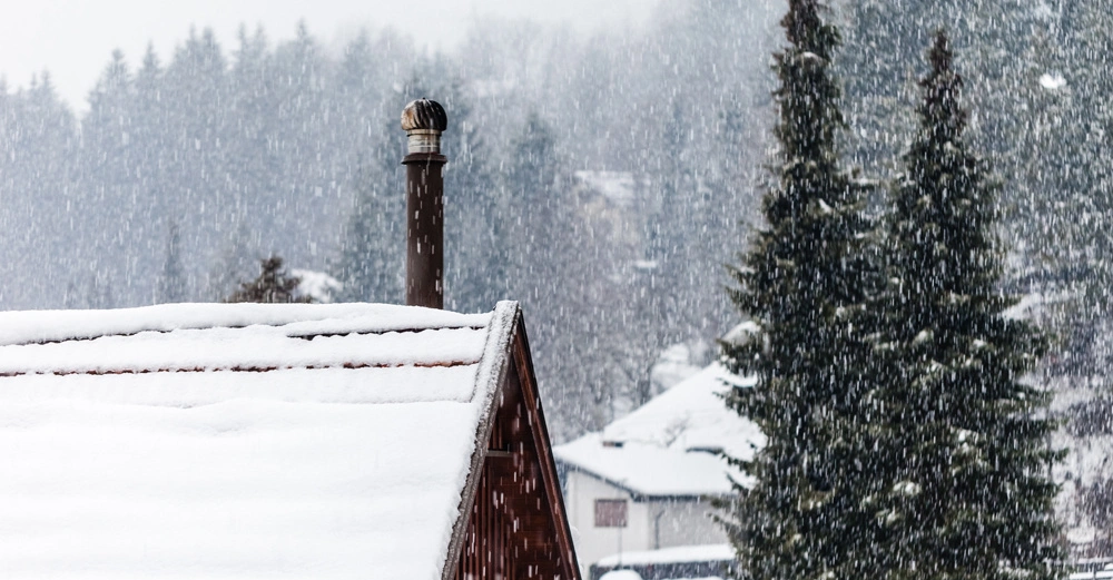 Ein schneebedecktes Dach hat einen Schornstein, aus dem kein Rauch steigt. Der Hintergrund zeigt einen verschneiten Wald und einige Häuser in einem Winterlandschaft. Es schneit stark.