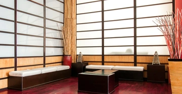 Sitzbänke mit weißen Kissen stehen in einem Raum mit Shoji-Trennwänden und Bambusdeko rote Holzböden ein Glastisch in der Mitte Laternen und hohen Vasen in den Ecken.