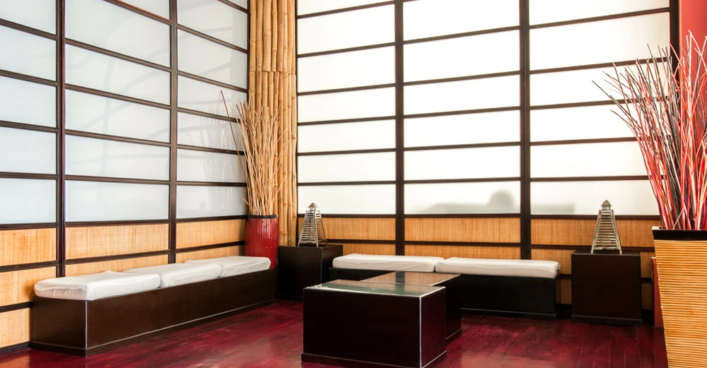 Sitzbänke mit weißen Kissen stehen in einem Raum mit Shoji-Trennwänden und Bambusdeko rote Holzböden ein Glastisch in der Mitte Laternen und hohen Vasen in den Ecken.