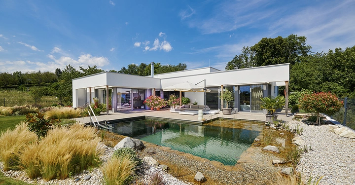 Galerie-Bild 3: Großbildaufnahme eines einstöckigen Holz-Fertigbaus von Magnum Vollholzdesign. Weiße Fassade, Fensterfronten, große Terrasse, Schwimmteich vor dem Haus. Sehr naturbelassene Atmosphäre