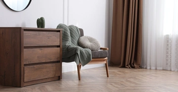Ein brauner Holzschrank steht neben einem grauen Sessel der mit einer grauen Decke und einem runden Kissen dekoriert ist. Im Hintergrund sind braune Vorhänge und ein Fenster zu sehen.