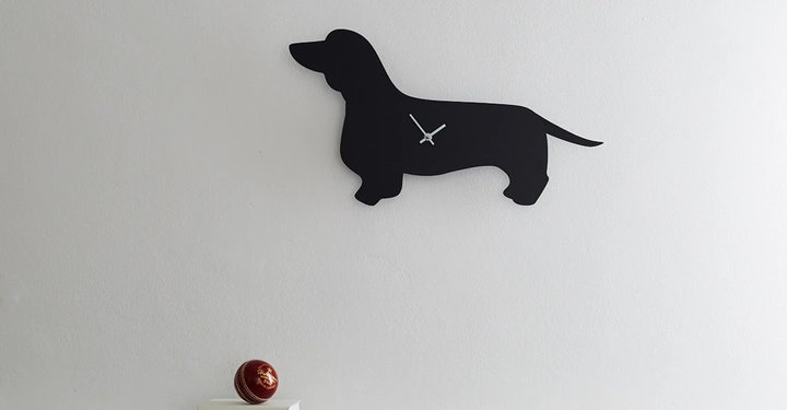 Galerie-Bild 10: Schwarze Wanduhr geformt wie ein Dackel an einer weißen Wand über einem Marmortisch auf dem ein roter Ball auf einem Stapel weißer Bücher liegt