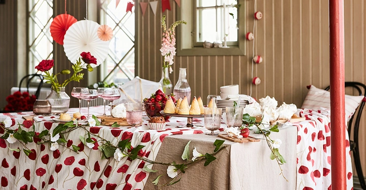 Ein festlich gedeckter Tisch mit Vasen Blumen und Glasgeschirr ist in einem hellen Raum mit Holzwänden und großer Fensterfront in einer fröhlich dekorierten Umgebung für eine Feier vorbereitet.
