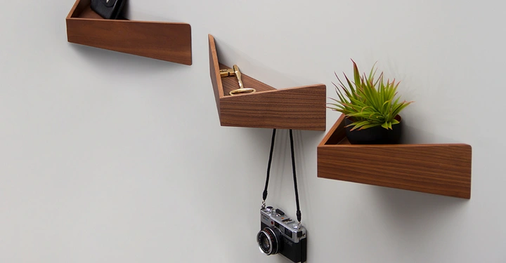 Galerie-Bild 3: Drei kleine, spitz zulaufende Walnussholz-Regale von WOODENDOT mit Ablagefläche und Haken an der Unterseite, am Regal in der Mitte baumelt eine Kamera