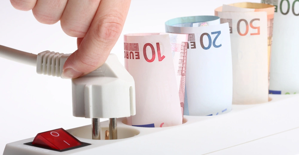 Hand steckt Stecker in Steckdosenleiste umgeben von aufgerollten Euro-Geldscheinen in verschiedenen Wertigkeiten im hellen Licht.