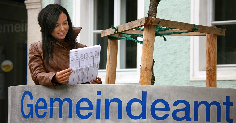 Eine Frau liest ein Dokument in einer Ledermantel im Freien vor einem Gebäude mit der Aufschrift Gemeindeamt. Das Gebäude hat eine grüne Fassade und Fenster.