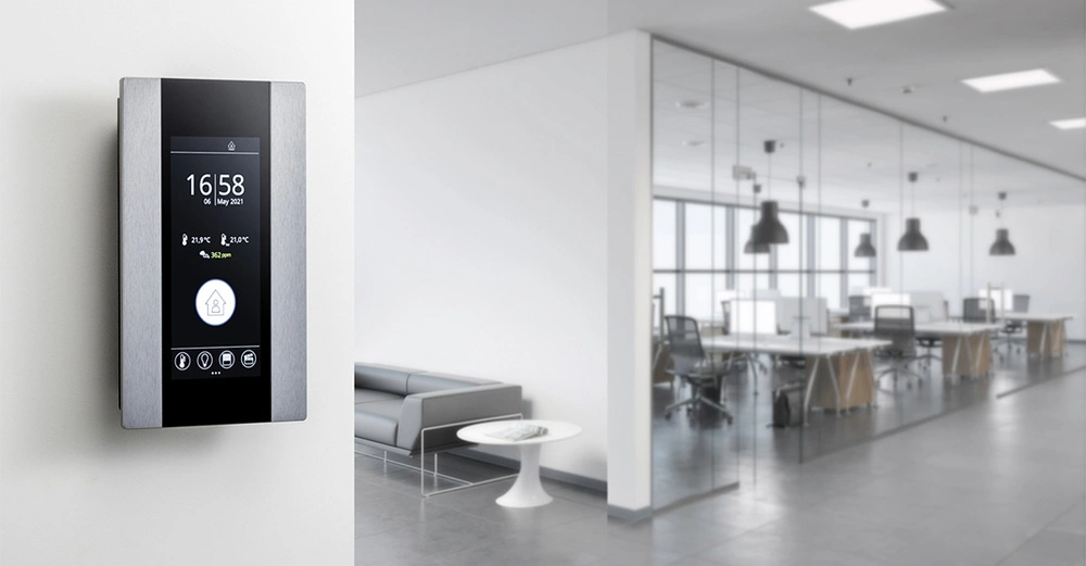 Ein Wand-Touchscreen zeigt 16 58 06 May 2021 und Raumklimadaten an Monitor links situiert In einem modernen Büro mit Glastrennwänden Bürotischen und Stühlen im Hintergrund