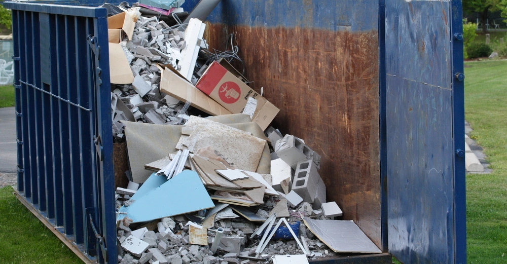 Ein großer blauer Müllcontainer steht auf einer Wiese und einer Straße; er ist voll mit diversen Bauschutt wie Ziegelsteine, Kartons und Holzabfälle.