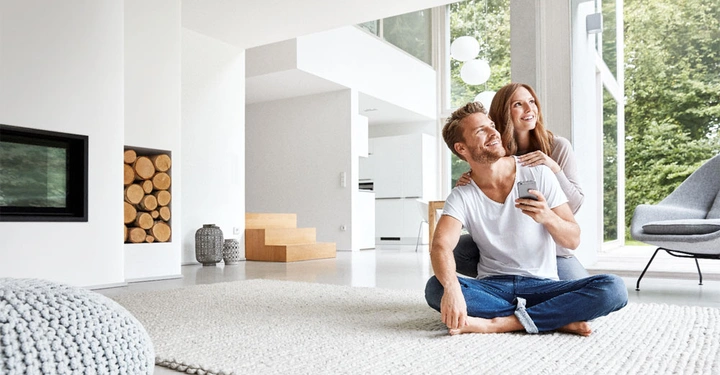 Ein Mann und eine Frau sitzen lächelnd auf einem hellen Teppich in einem modernen Wohnzimmer mit großen Fenstern der Mann hält ein Smartphone in einer Hand Holzscheite sind neben einem eingebauten Kamin gestapelt.