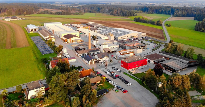 Luftaufnahme einer großen Industrieanlage arbeitet in einer ländlichen Umgebung. Felder, Wälder und eine Straße umgeben die Fabrik und mehrere Parkplätze und Gebäude befinden sich auf dem Gelände.