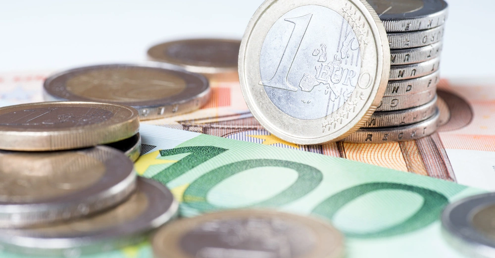 Münzen liegen auf gestapelten Banknoten Euro in verschiedenen Nennwerten auf einem Tisch. Eine Ein-Euromünze ist prominent sichtbar im Zentrum der Szene.