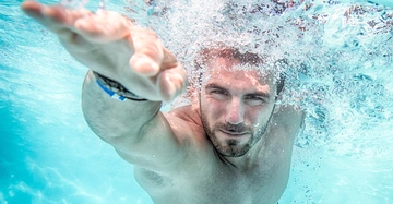 Mann schwimmt unter Wasser und streckt einen Arm nach vorne aus. Blasen umgeben sein Gesicht im klaren blauen Poolwasser.