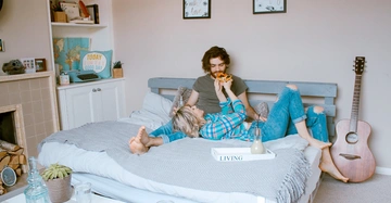 Personen essen Pizza auf Bett gemütliches Schlafzimmer mit Gitarre Bücherregalen Pflanzen und Dekorationen Im Hintergrund ein Schild mit "TODAY is going to be a GOOD DAY" Text auf Tablett "LIVING".