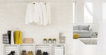 Weiße Hemden hängen an einem Ast an einer weißen Backsteinwand über Holzkisten mit Schuhen und Einkaufstüten im modernen, hellen Wohnzimmer mit Sofa und großen Fenstern.