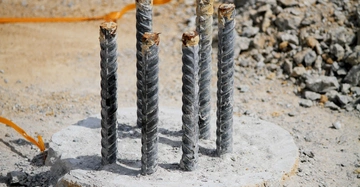 Nahaufnahme eines Betonpfahls mit sechs Eisenverstrebungen, dessen Ende auf einer Baustelle aus dem staubigen Boden ragt.
