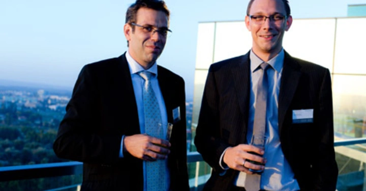 Galerie-Bild 6: Zwei Männer in Anzügen und Krawatten halten Getränke und stehen auf einer Terrasse mit einer Glasbrüstung bei Tageslicht im Freien. Eine Stadtlandschaft ist im Hintergrund sichtbar.