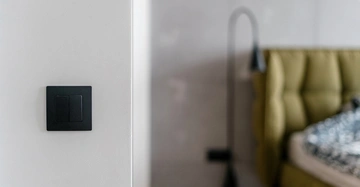 Schwarzer Wandschalter angebracht an weißer Wand links Schlafzimmereinrichtung mit grünem gepolstertem Kopfteil und Bett rechts im Hintergrund