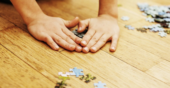 Kinderhände legen ein Puzzle auf einem Holzboden zusammen während einzelne Puzzleteile verstreut um sie herumliegen.