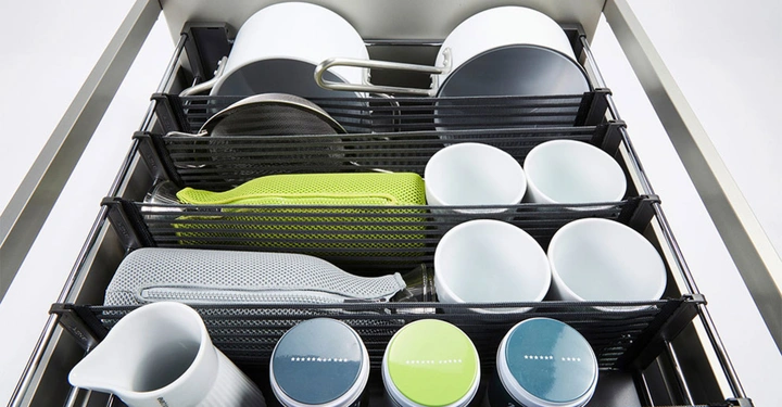 Geschirr und Küchenutensilien liegen ordentlich in einer Schublade in einem Netzorganizer. Der Kontext ist eine moderne Küche mit Metallrahmen.