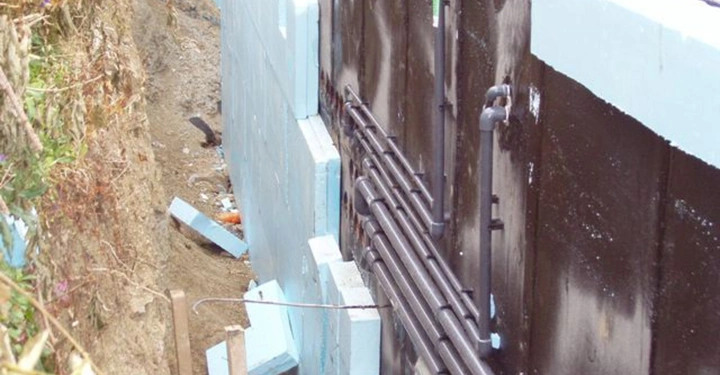 Galerie-Bild 22: Isolierung wird an der Außenseite eines Gebäudefundaments angebracht mehrere Rohre verlaufen entlang der Wand in einem Graben mit Erde und Bauschutt darunter.