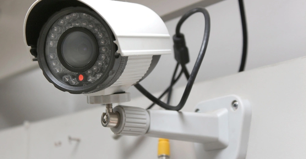 Überwachungskamera hängt an einer Wandhalterung und ist an zwei Kabel angeschlossen Umgebung ist eine schlicht eingerichtete Innenraumumgebung mit hellen Wänden.