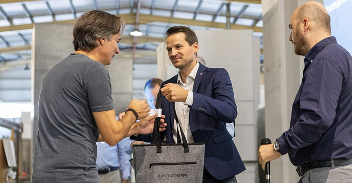 drei Männer in dunklen Oberteilen stehen in großer Fertigungshalle. Mann in der Mitte übergibt ein Fertigungsteil an den linksstehenden. 