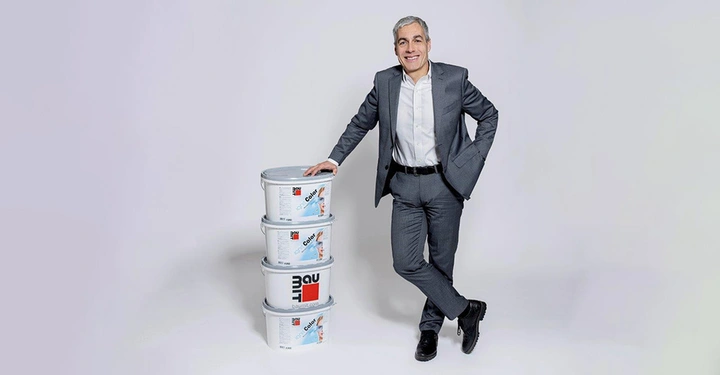Mann in grauem Anzug lehnt an einem Stapel von vier Eimern mit der Aufschrift "baumit Color" in einem hellen Studio.