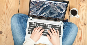 Person tippt auf Laptop mit Kartenansicht auf dem Bildschirm sitzend auf Holzfußboden neben Tasse Kaffee.