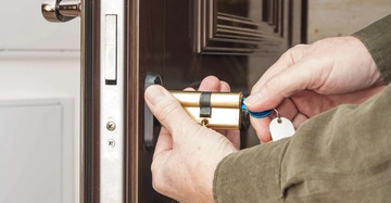 Hände ersetzen ein Türschloss in einem Hauseingang indem sie einen Zylinder mit einem blauen Schlüssel befestigen im Hintergrund ist eine Holztür und ein Teil eines weißen Türrahmens sichtbar