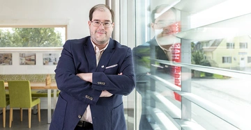Mann in Anzug steht mit verschränkten Armen vor einer Glaswand in einem modern eingerichteten Raum und lächelt leicht die Umgebung zeigt Tische Stühle und Fenster mit Blick nach draußen