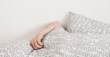 Ein Arm ragt unter einer Bettdecke hervor auf einem Bett mit weiß-grauer wellenförmiger Bettwäsche in einem weißen Raum.