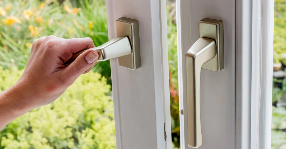 Eine Hand öffnet ein Fenstergriff Die Hand hält einen goldenen Fenstergriff in einem hellen Raum Im Hintergrund ist grüner Garten mit Blumen zu sehen
