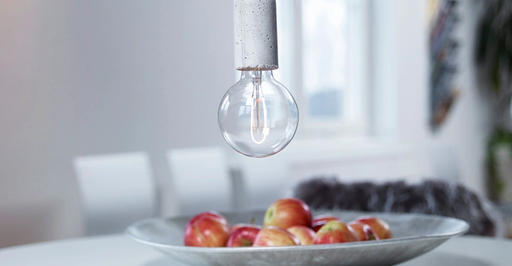 Galerie-Bild 2: Glühbirne hängt von der Decke schwebend über einer Metallschale mit Äpfeln in einem hellen Raum mit unscharfen weißen Stühlen und einem Fenster im Hintergrund.