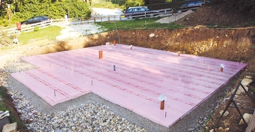 Rosa Dämmplatten liegen auf einem vorbereiteten Bauplatz während des Hausbaus mit Schotterumgebung und geparkten Autos auf einer Schotterstraße im Hintergrund.